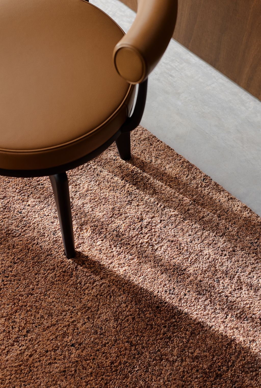 A modern brown chair with a round cushioned seat and curved armrest rests on the Meadow Rug - Brick by Armadillo&amp;Co, casting a shadow across the rug’s textured surface and the nearby light gray floor.