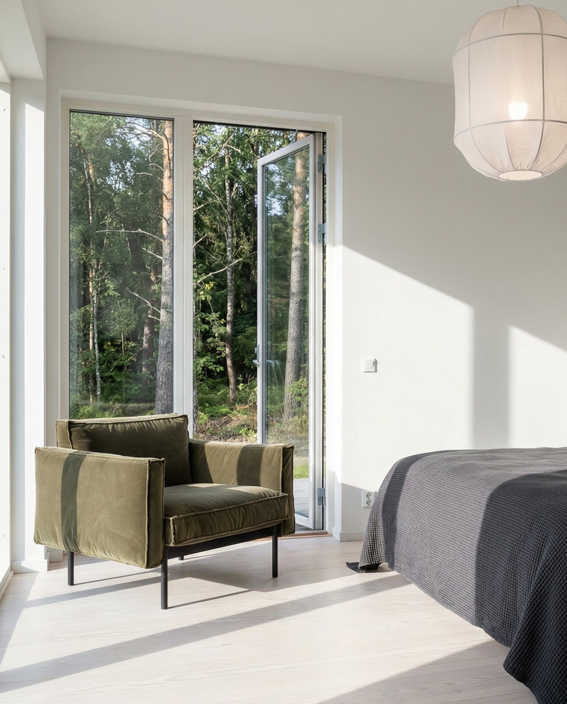 A modern bedroom with a large windowed door overlooking a green forest. Sunlight fills the space, which features Wooden Stories' Attendant Lounge Chair | Fabric, a gray bedspread, light wood floors, and a white pendant lamp.