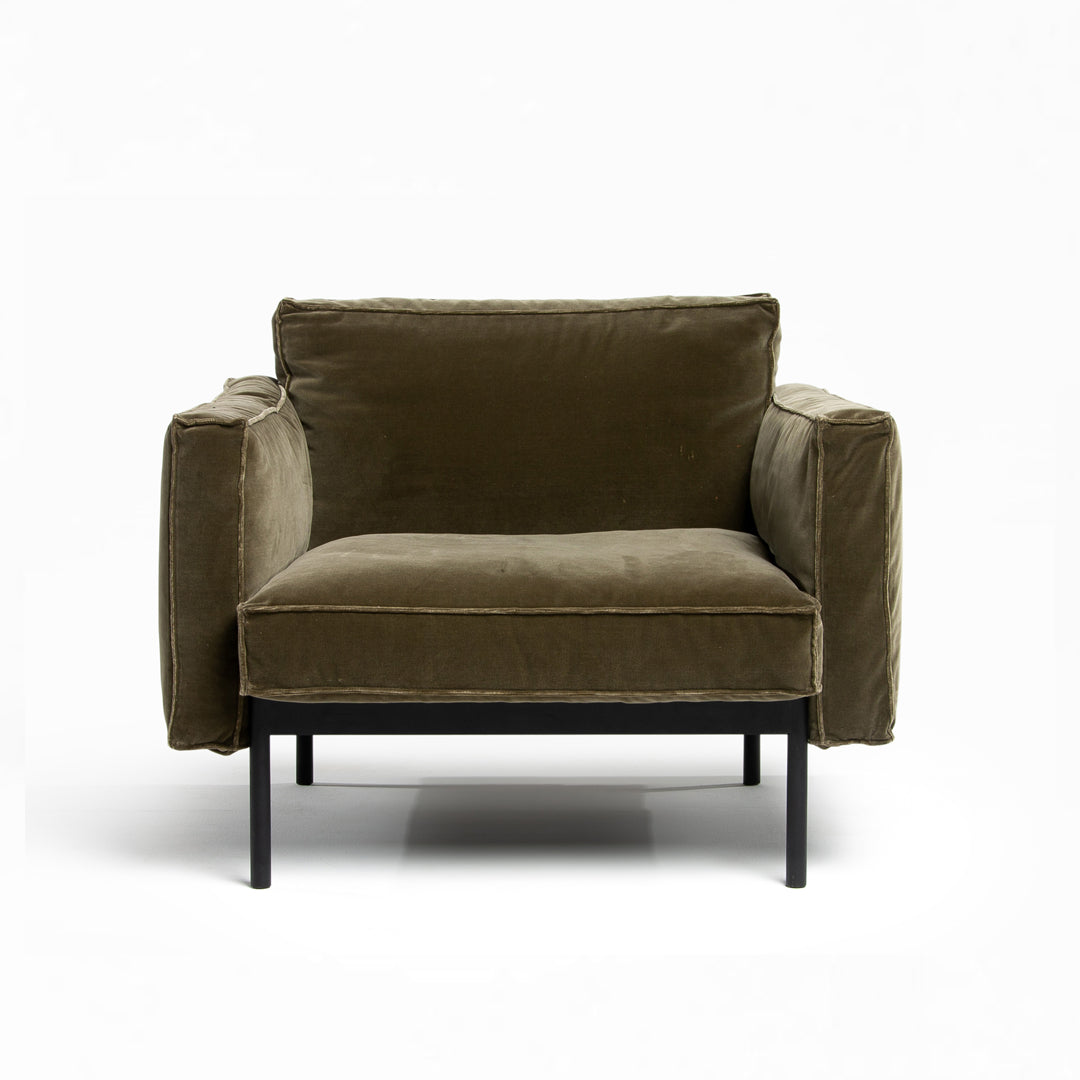 The Attendant Lounge Chair by Wooden Stories features plush, boxy cushions in modern olive green fabric and slim black metal legs, shown here against a plain white background.