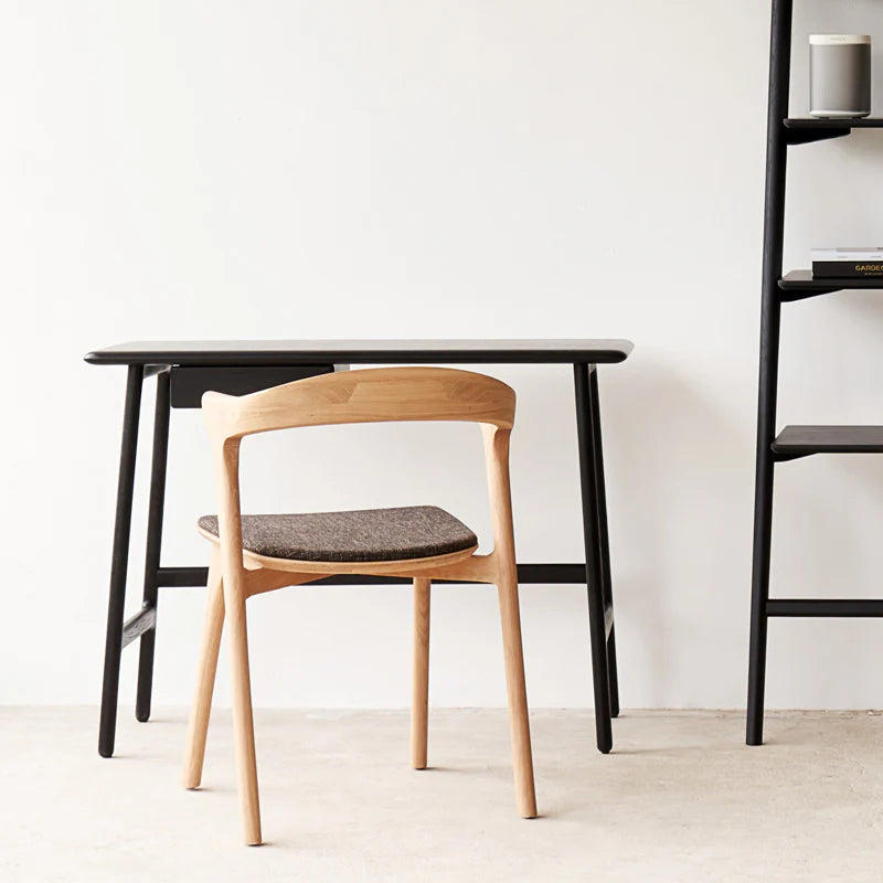 A minimalist home office featuring the Wooden Stories Author Desk in black oak, a light wooden chair, and a black shelving unit with a candle and books, all set against a plain white wall.