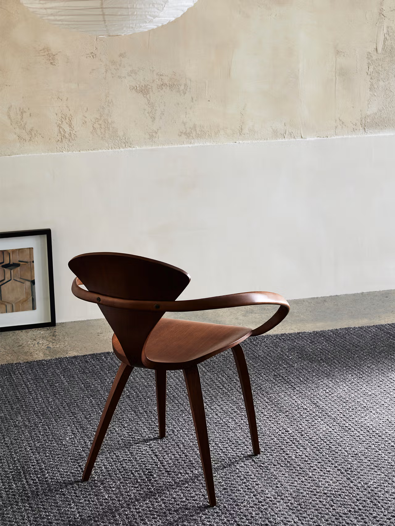 A wooden chair with curved armrests sits on the Sherpa Rug in Charcoal by Armadillo&Co in a minimalist room. A framed picture leans against the wall, and a paper lantern hangs from the ceiling above.