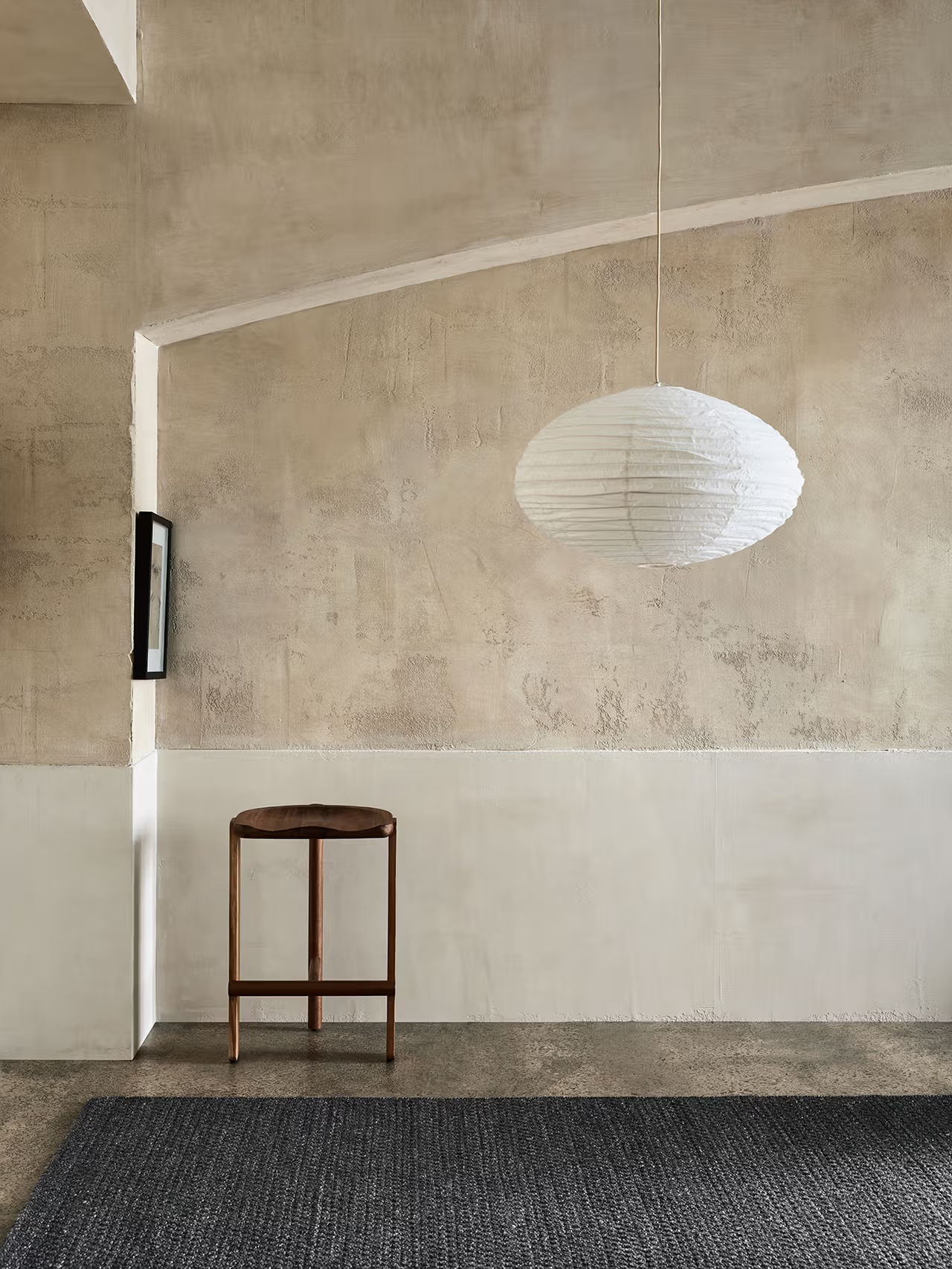 A minimalist room with textured beige walls features the Sherpa Rug in Charcoal by Armadillo&Co, accompanied by a small wooden table and a large white paper lantern pendant light hanging from the ceiling.