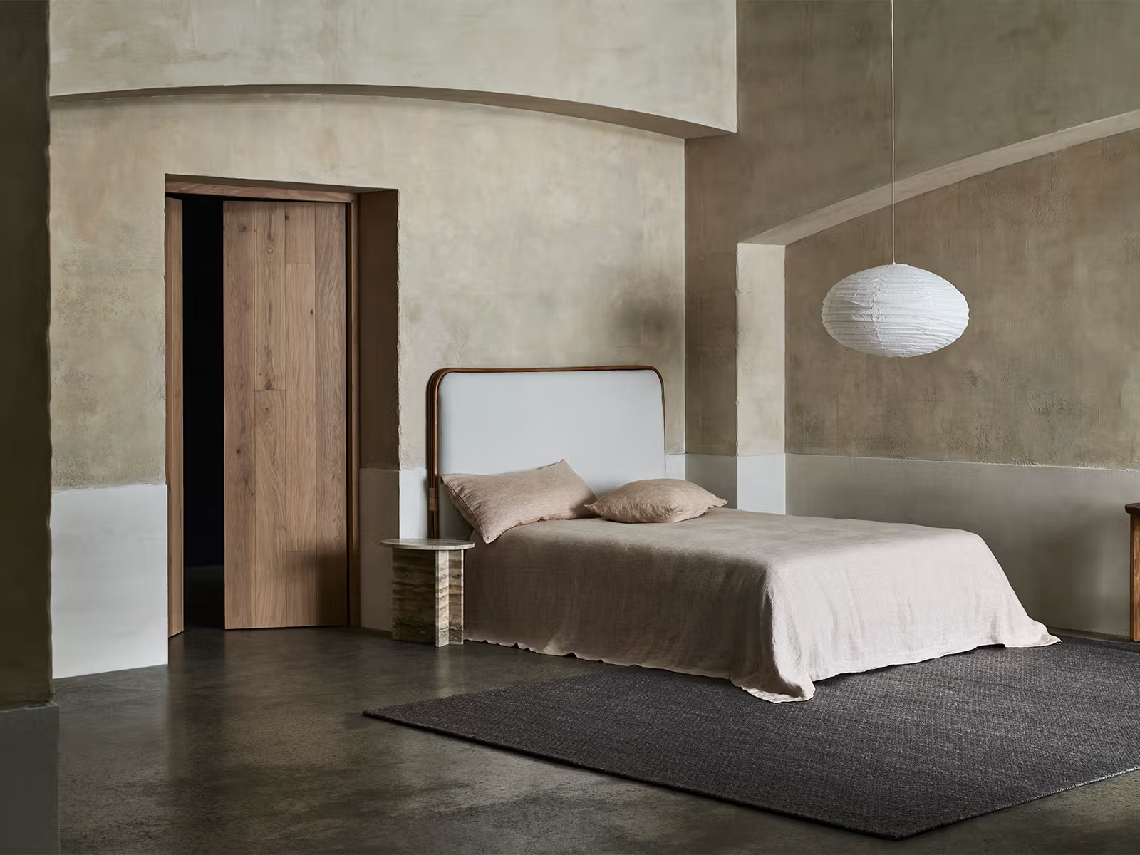 A minimalist bedroom featuring a single bed with beige bedding, a round nightstand, and the Sherpa Rug in Charcoal by Armadillo&amp;Co adding soft gray texture underfoot. A white paper lantern pendant hangs overhead, while textured neutral walls complement the arched ceiling and wooden door.