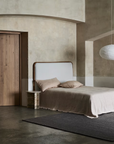 A minimalist bedroom featuring a single bed with beige bedding, a round nightstand, and the Sherpa Rug in Charcoal by Armadillo&Co adding soft gray texture underfoot. A white paper lantern pendant hangs overhead, while textured neutral walls complement the arched ceiling and wooden door.