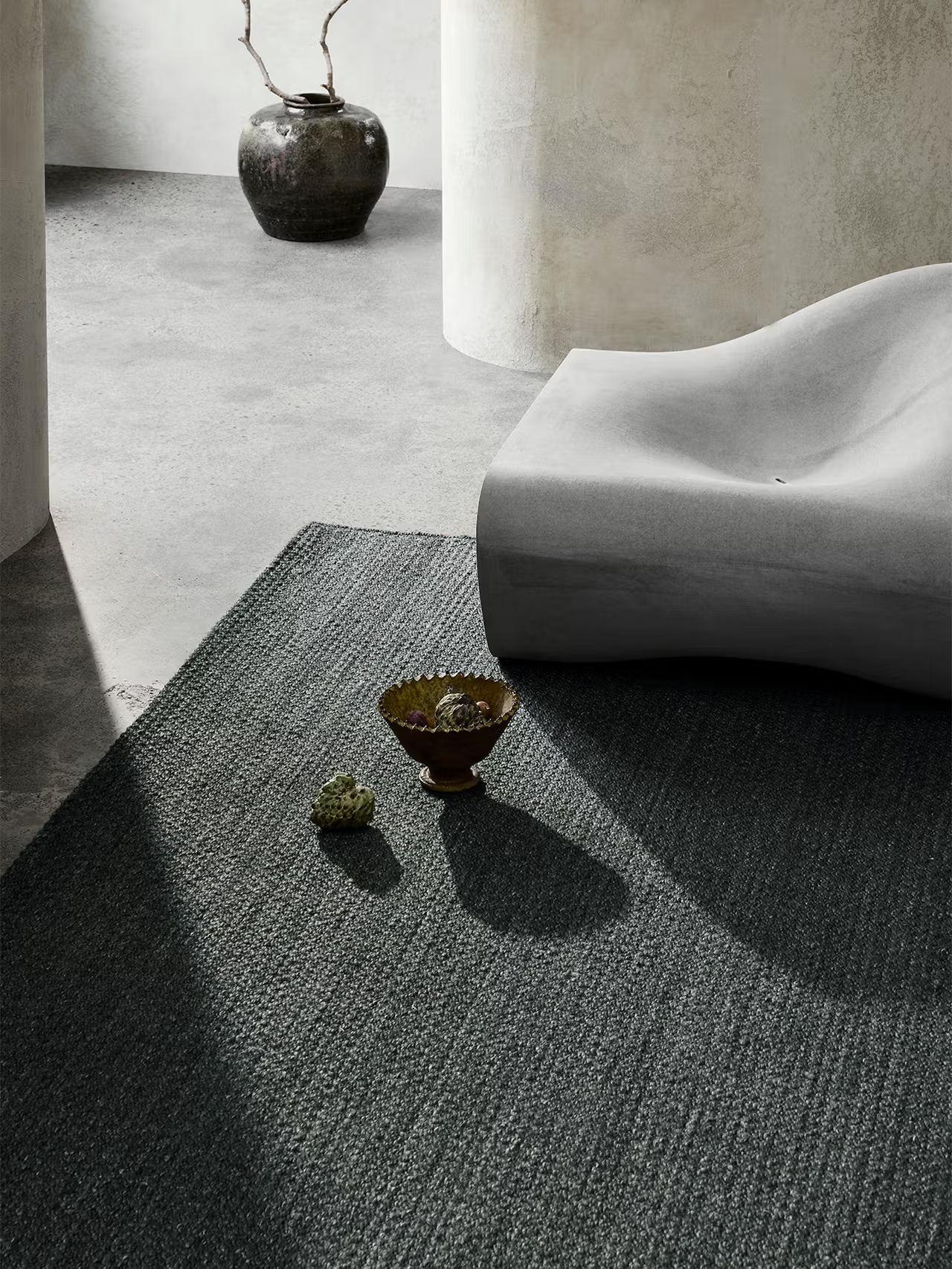 A modern minimalist room featuring the Sherpa Rug – Kelp by Armadillo&amp;Co, a gray hand-woven wool rug paired with a sculptural gray lounge chair, a ceramic vase holding branches, a decorative bowl, and two small stones placed atop the rug. Sunlight streams in, casting gentle shadows across the space.