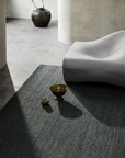A modern minimalist room featuring the Sherpa Rug – Kelp by Armadillo&Co, a gray hand-woven wool rug paired with a sculptural gray lounge chair, a ceramic vase holding branches, a decorative bowl, and two small stones placed atop the rug. Sunlight streams in, casting gentle shadows across the space.