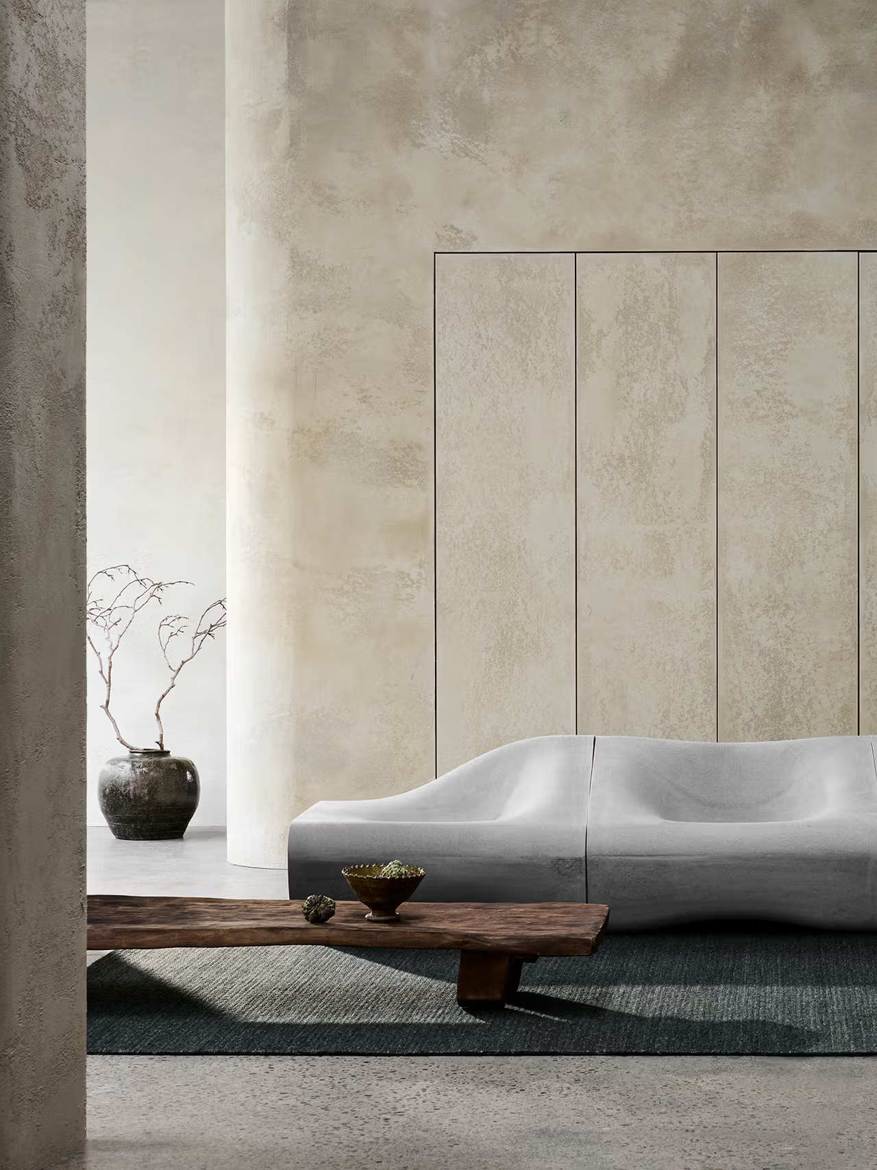 Minimalist living room featuring a sculptural gray sofa, a low wooden coffee table, the Sherpa Rug in Kelp by Armadillo&Co, and a ceramic vase with bare branches set against textured beige walls and columns.