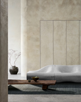 Minimalist living room featuring a sculptural gray sofa, a low wooden coffee table, the Sherpa Rug in Kelp by Armadillo&Co, and a ceramic vase with bare branches set against textured beige walls and columns.