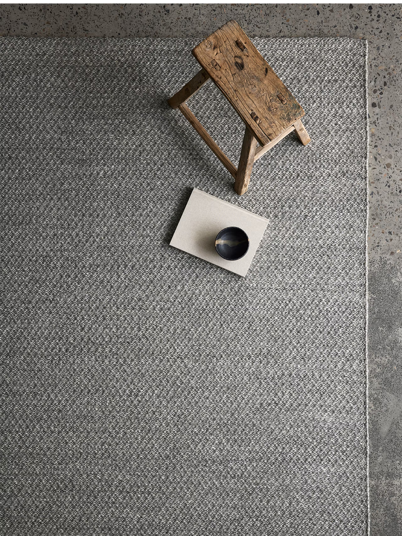 A small wooden stool and a beige book topped with a dark bowl sit on the Armadillo&Co Sherpa Rug in Pumice, with part of a speckled concrete floor visible at the edge.