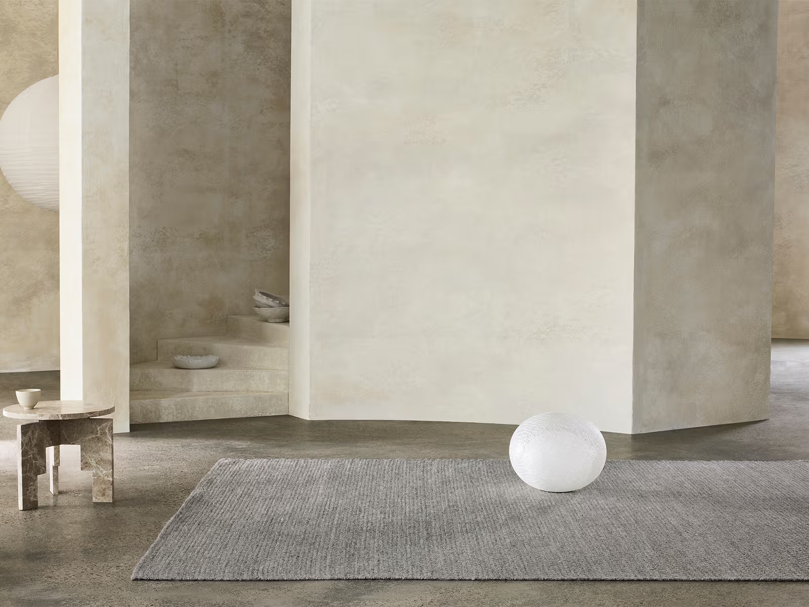 Minimalist room with cream walls, an Armadillo&amp;Co Sherpa Rug in Pumice, a white spherical object, a small marble side table with a cup, and some decorative bowls on stone steps in the background.