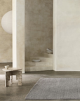 Minimalist room with neutral tones, featuring a marble side table with a cup, the Armadillo&Co Sherpa Rug in Pumice handwoven on the concrete floor, sculptural bowls on curved stairs, and a large lamp partially visible on the left.