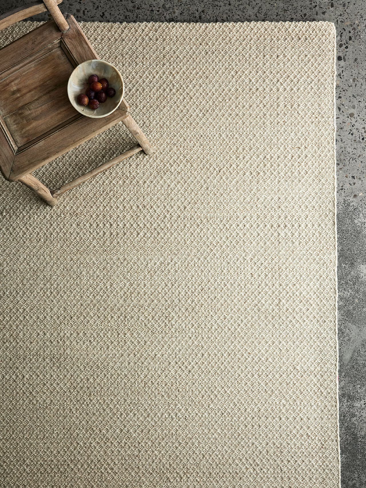 The Armadillo&Co Sherpa Rug in Sand, a Fair Trade handwoven wool piece, rests on a gray textured floor, while a rustic wooden chair in the corner holds a bowl of grapes.