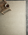 The Armadillo&Co Sherpa Rug in Sand, a Fair Trade handwoven wool piece, rests on a gray textured floor, while a rustic wooden chair in the corner holds a bowl of grapes.