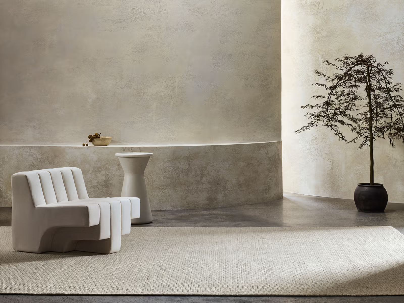 A minimalist room featuring a modern white lounge chair, a small side table with a bowl, a large potted tree, and a neutral textured wall, anchored by the Sherpa Rug - Sand from Armadillo&amp;Co—a handwoven light beige wool rug laid over polished concrete flooring.