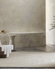 A minimalist room featuring a modern white lounge chair, a small side table with a bowl, a large potted tree, and a neutral textured wall, anchored by the Sherpa Rug - Sand from Armadillo&Co—a handwoven light beige wool rug laid over polished concrete flooring.