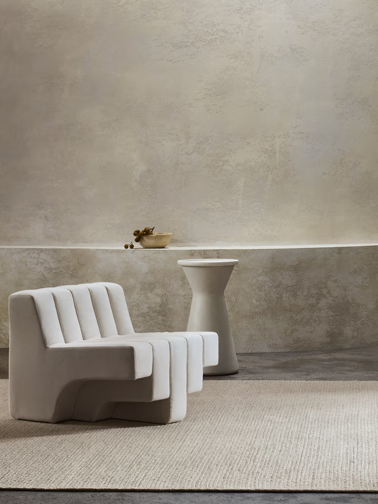 Minimalist room featuring a sculptural white chair, the Armadillo&Co Sherpa Rug in Sand, a round pedestal table, and a shallow bowl with dried branches against a neutral plaster wall.