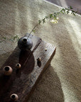 A rustic wooden coffee table holds a dark vase with a leafy branch and small clay pots, set on the Armadillo&Co Crest Rug in Mica with a herringbone pattern, as sunlight streams across the serene scene.