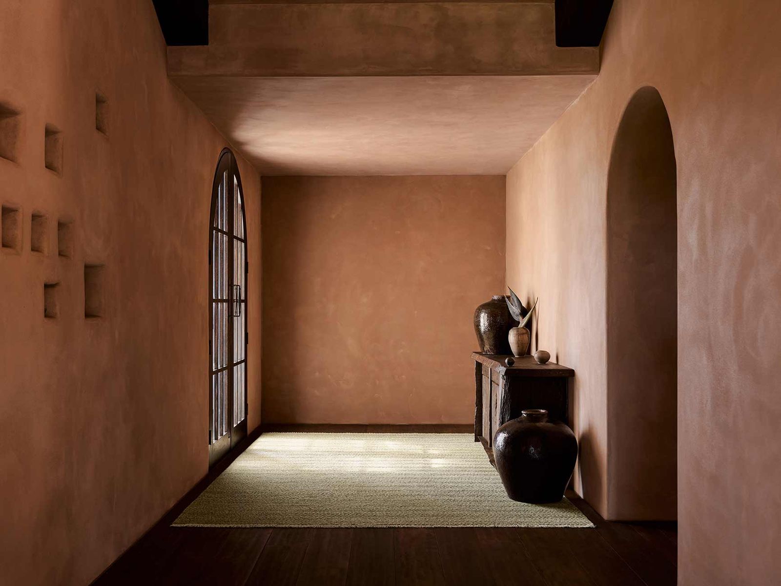 A minimalist hallway with earthy walls, arched doorways, and a console table with vases and dried plants features the Crest Rug - Mica by Armadillo&amp;Co, adding subtle texture and calm to the naturally lit space.