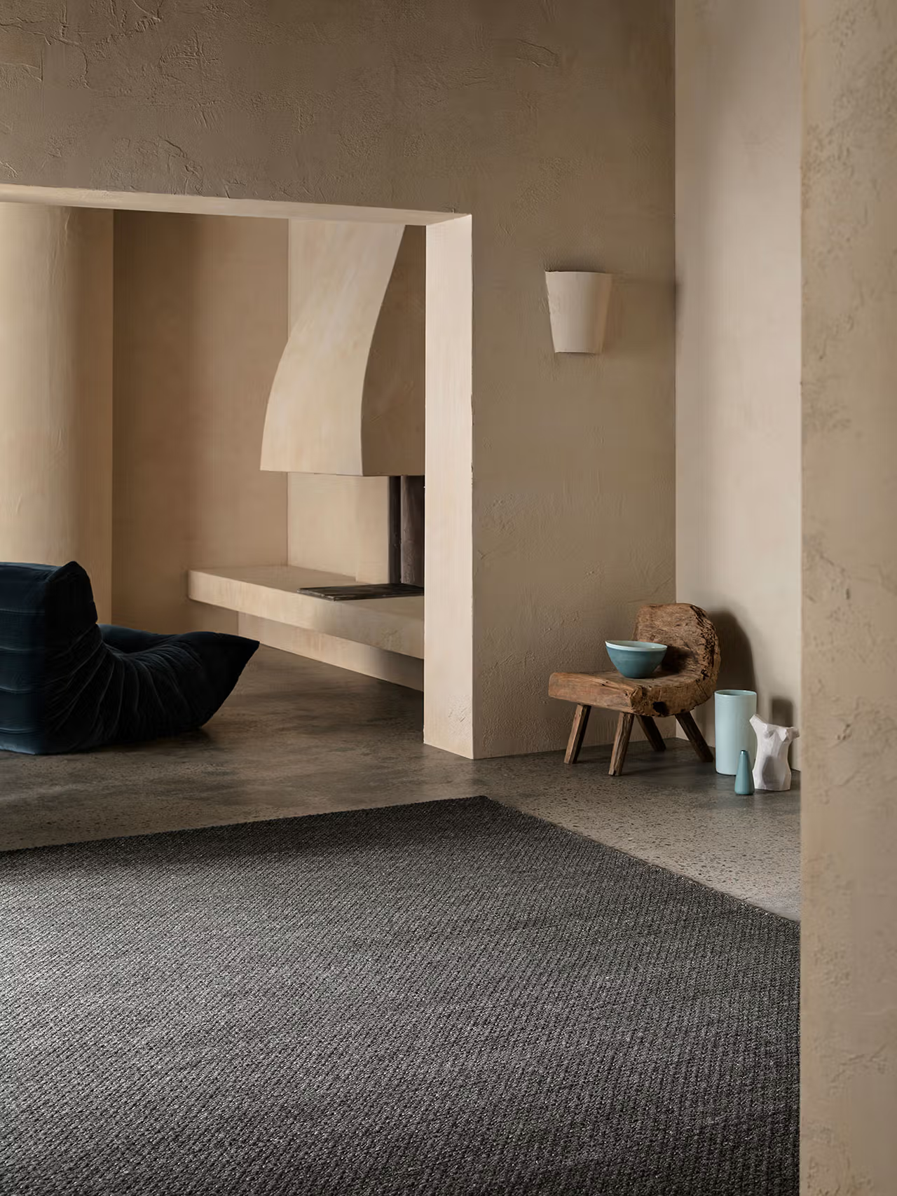 A minimalist living space with neutral walls features a modern black lounge chair and a rustic wooden stool topped with a blue bowl, accompanied by small decorative vases. The room is grounded by the Sherpa Rug in Charcoal from Armadillo&amp;Co, which adds subtle texture beneath the furnishings. In the background, a fireplace is built into the cream-colored wall.
