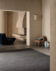 A minimalist living space with neutral walls features a modern black lounge chair and a rustic wooden stool topped with a blue bowl, accompanied by small decorative vases. The room is grounded by the Sherpa Rug in Charcoal from Armadillo&Co, which adds subtle texture beneath the furnishings. In the background, a fireplace is built into the cream-colored wall.