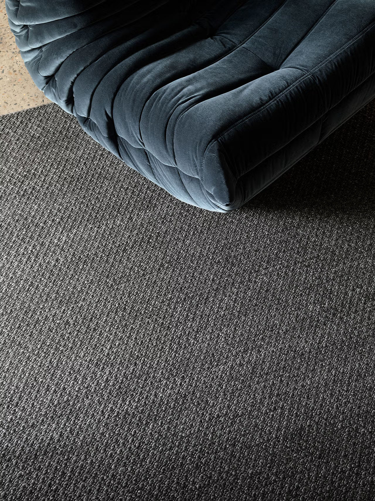 An elegant, dark velvet tufted chair rests atop a textured gray carpet, complemented by the subtle presence of the Armadillo&amp;Co Sherpa Rug in Charcoal, with a glimpse of speckled concrete floor visible in the top left corner.