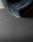 An elegant, dark velvet tufted chair rests atop a textured gray carpet, complemented by the subtle presence of the Armadillo&Co Sherpa Rug in Charcoal, with a glimpse of speckled concrete floor visible in the top left corner.