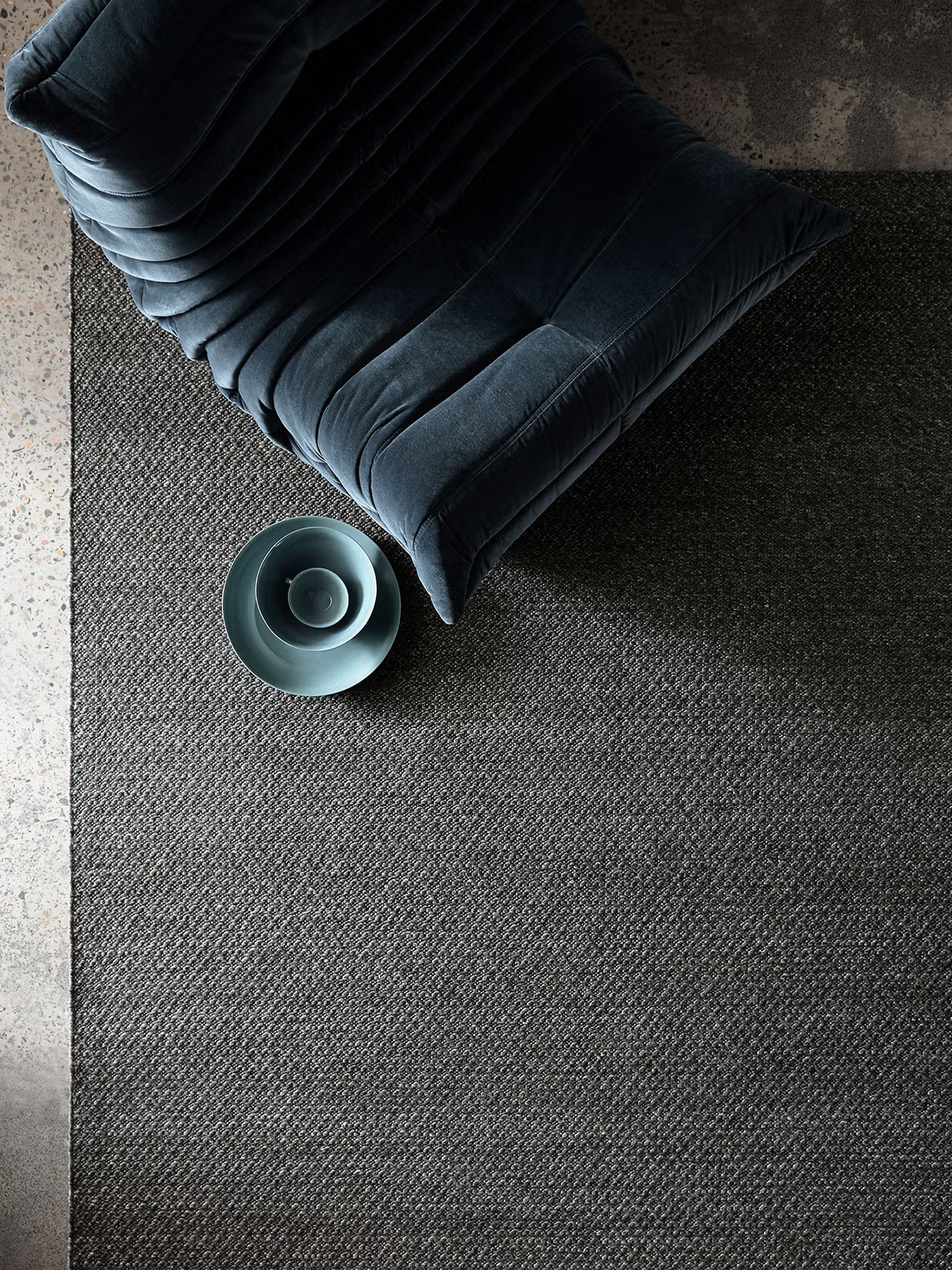 A plush chair in dark blue sits atop the Armadillo&amp;Co Sherpa Rug in Charcoal, next to a light blue bowl, all viewed from above. The scene is minimal and modern, illuminated by soft natural light.