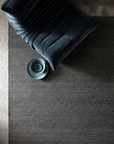 A plush chair in dark blue sits atop the Armadillo&Co Sherpa Rug in Charcoal, next to a light blue bowl, all viewed from above. The scene is minimal and modern, illuminated by soft natural light.