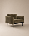 The Attendant Lounge Chair by Wooden Stories features olive green velvet, boxy cushions, and black metal legs, all expertly handcrafted. Shown here against a plain beige background.