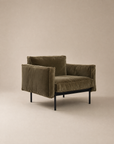 The Attendant Lounge Chair by Wooden Stories features olive green velvet, boxy cushions, and black metal legs, all expertly handcrafted. Shown here against a plain beige background.