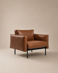 The Attendant Lounge Chair | Leather by Wooden Stories features brown leather upholstery, square cushions, wide armrests, and a black metal frame, set against a plain beige background.