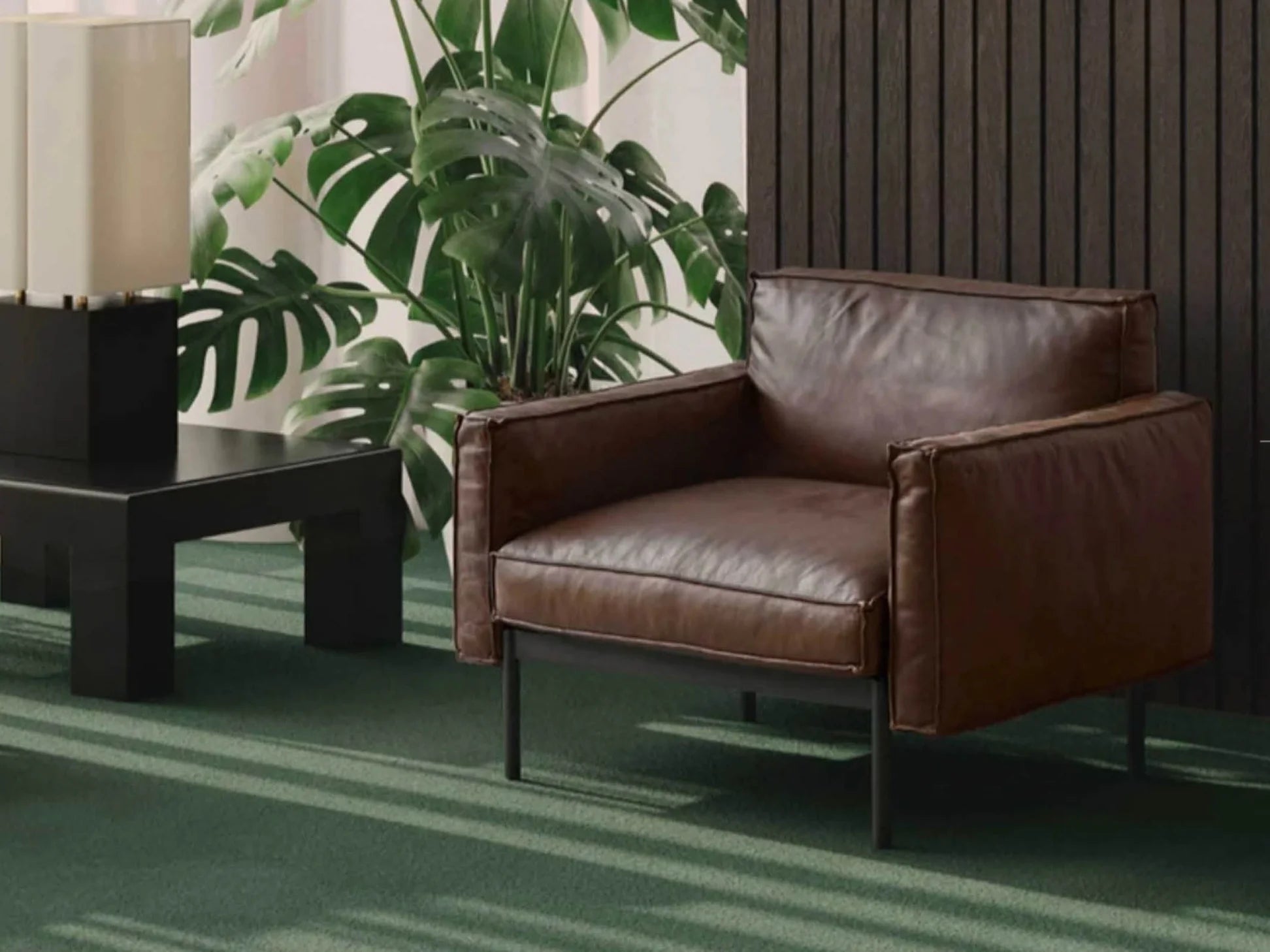 A modern room with the Wooden Stories Attendant Lounge Chair in brown leather featuring French seams, placed on a green carpet with a black coffee table, lamp, large green plants, and feather-padded panels on a wood-paneled wall.