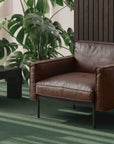 A modern room with the Wooden Stories Attendant Lounge Chair in brown leather featuring French seams, placed on a green carpet with a black coffee table, lamp, large green plants, and feather-padded panels on a wood-paneled wall.