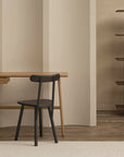A minimalist home office features the Author Desk in Light Oak by Wooden Stories and a black chair on a light rug. In the background, a wooden shelf with ceramic bowls and books rests against a beige wall.