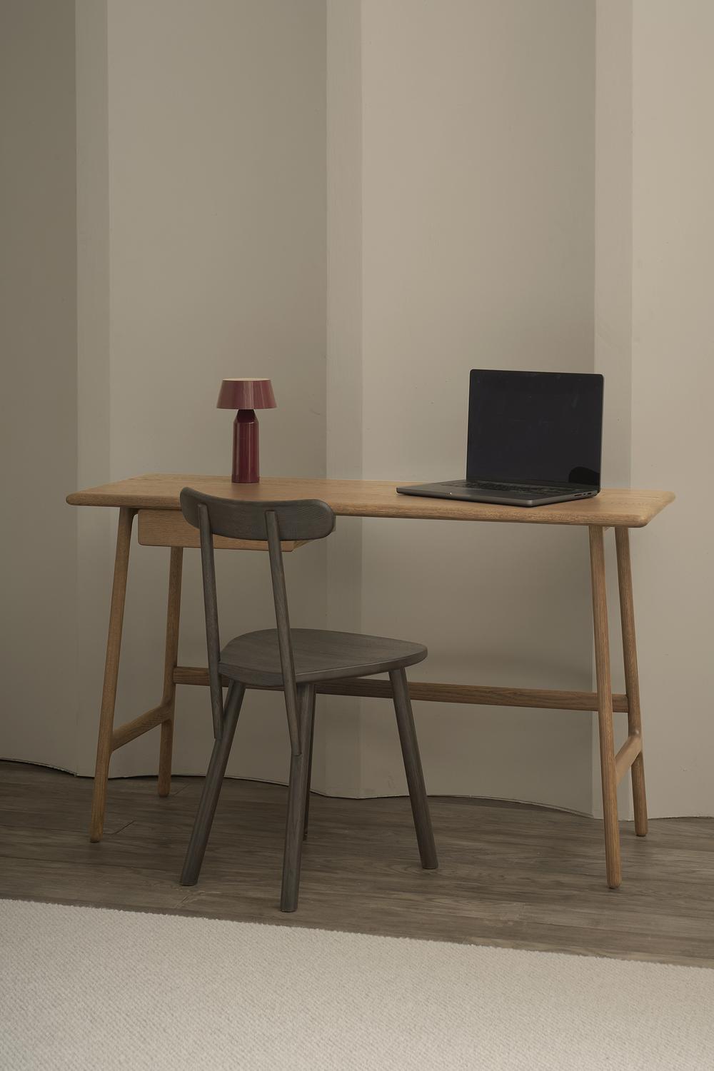 A minimalist home office highlights the Wooden Stories Author Desk in light oak, paired with a dark wooden chair and red lamp, all set against a beige wall with a light rug—perfect for a stylish workspace.