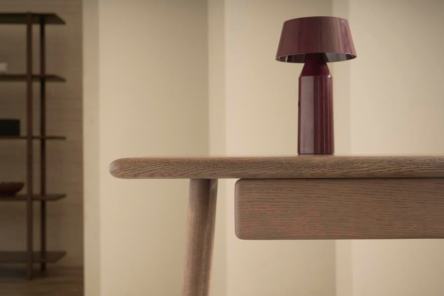 The Author Desk - Light Oak by Wooden Stories, featuring rounded edges, displays a modern, dark red table lamp. Neutral walls and a wooden shelving unit create an ideal Scandinavian desk setup for your home office.