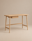 The Wooden Stories Author Desk in light oak features angled legs and a single central drawer, shown against a neutral beige background.