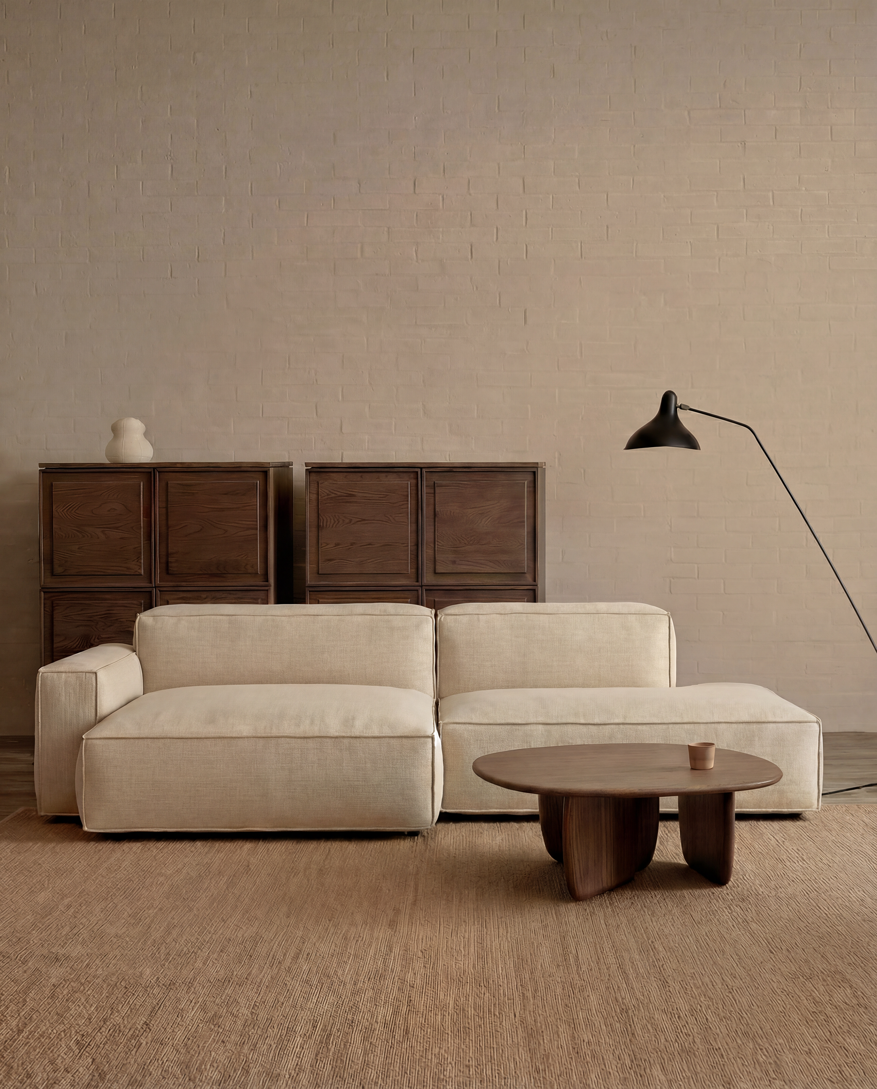 Minimalist living room with a beige Baker Modular Sofa | Fabric • M4 by Wooden Stories, round wooden coffee table, tall dark wood cabinets, floor lamp, and textured rug against a light brick wall.
