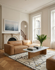A bright living room with the Baker Modular Sofa | Leather • M1 by Wooden Stories in tan leather, a marble coffee table, books, a vase of green leaves, a patterned rug, large windows, a white fireplace, and framed ocean artwork above the mantel.