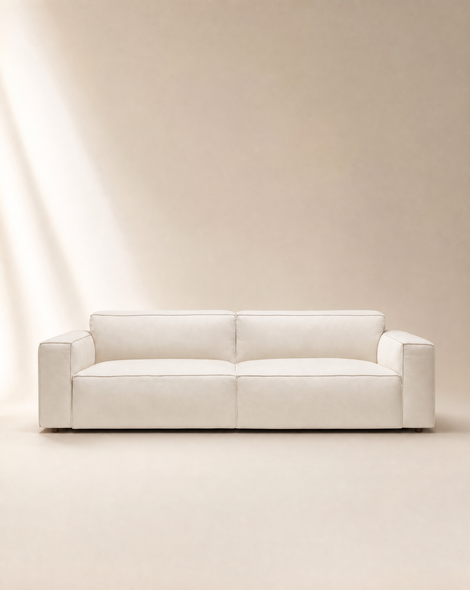 A modern Baker Sofa | Fabric by Wooden Stories, upholstered in cream fabric with wide armrests and a minimalist Danish design, features plush feather filling and is set against a neutral beige backdrop illuminated softly from the left.