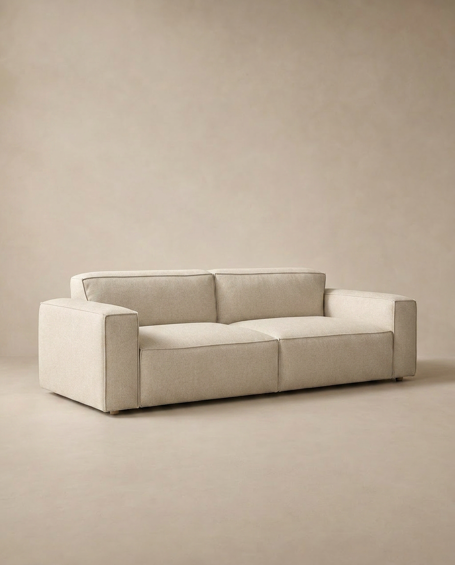 The Baker Sofa | Fabric by Wooden Stories features a modern, low-profile Danish design with feather filling, wide armrests, and clean lines, set against a matching beige background.