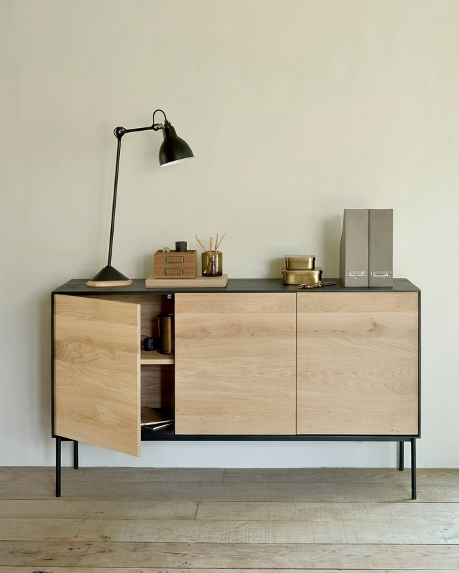 The Blackbird Sideboard by Ethnicraft, crafted from solid European oak and accented with sleek black legs, is styled with a black desk lamp, office supplies, and two folders arranged on its surface. One of its cabinet doors is slightly ajar, offering a glimpse of the items stored inside. This modern piece sits elegantly on a light wooden floor.
