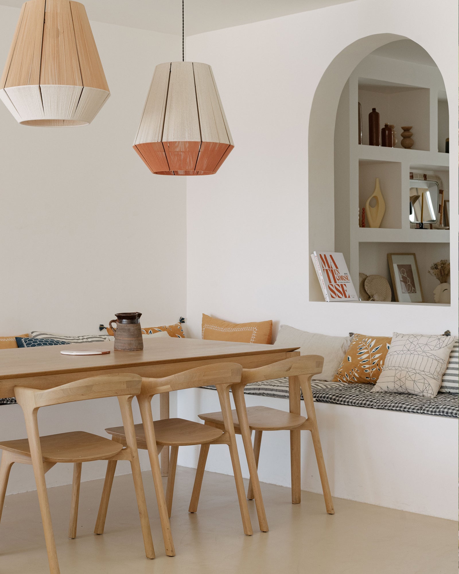 Minimalist dining area featuring the Ethnicraft Bok Dining Table in oak, pendant lamps, and a built-in bench with patterned and solid cushions. An arched wall niche displays books and decor in warm, neutral hues.