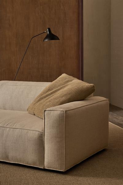 The Baker Modular Sofa | Fabric • M9 by Wooden Stories, in modern beige, features a large brown cushion and sits on a textured rug beneath a black minimalist wall lamp, set against a brown wooden panel and taupe wall.
