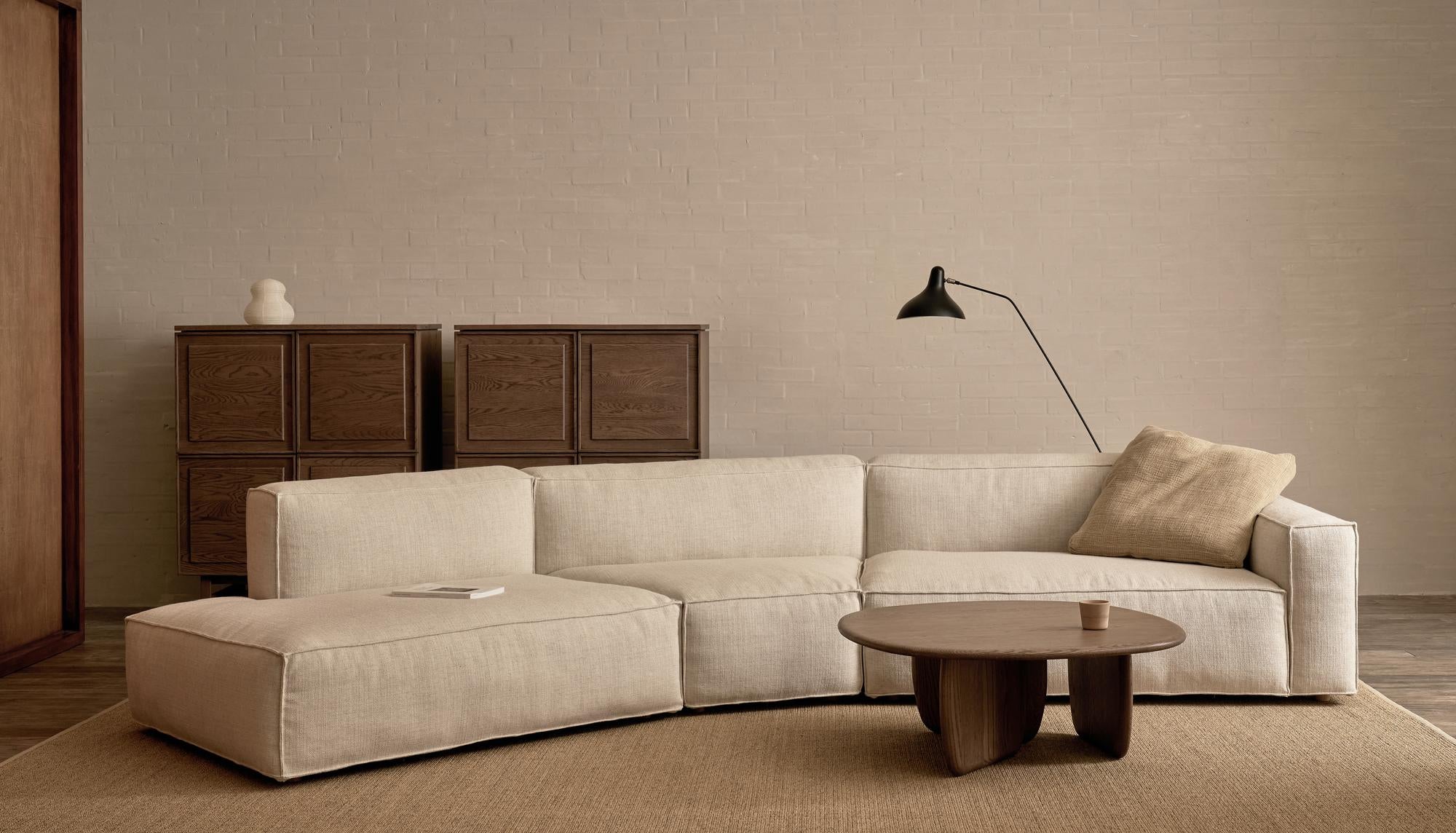 A modern living room showcases the Baker Modular Sofa | Fabric • M9 by Wooden Stories in beige, paired with a light brown cushion, round wooden coffee table, two wooden cabinets, floor lamp, and minimalist decor against a neutral brick wall.