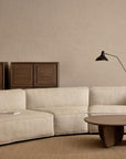 A modern living room showcases the Baker Modular Sofa | Fabric • M9 by Wooden Stories in beige, paired with a light brown cushion, round wooden coffee table, two wooden cabinets, floor lamp, and minimalist decor against a neutral brick wall.