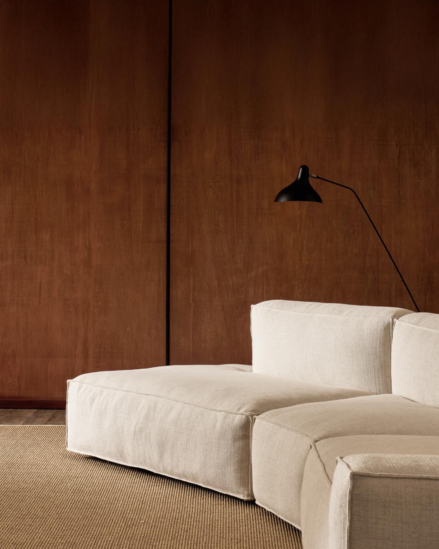 A modern, minimalist living room showcases the customizable Baker Modular Sofa | Fabric • M9 by Wooden Stories on a textured rug, set against a smooth brown wooden wall with a black adjustable wall-mounted lamp above.