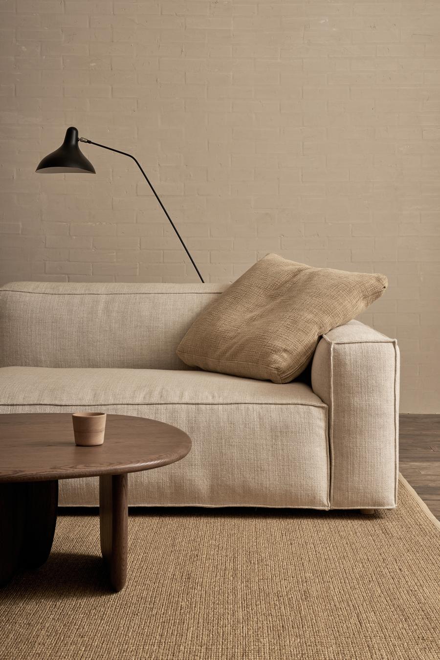 A minimalist living room features the Baker Modular Sofa | Fabric • M9 by Wooden Stories in beige, a textured tan cushion, wooden coffee table with a small cup, woven rug, and a black wall lamp set against a neutral brick wall.