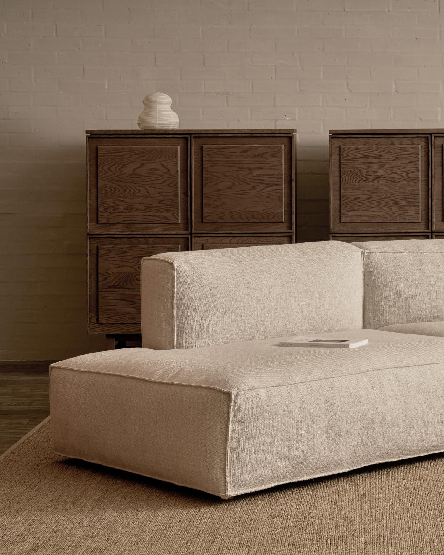 A minimalist living room features the Baker Modular Sofa | Fabric • M9 by Wooden Stories in beige, a book on its seat, wooden cabinets with paneled doors behind, a decorative vase on top, and a neutral-toned brick wall.