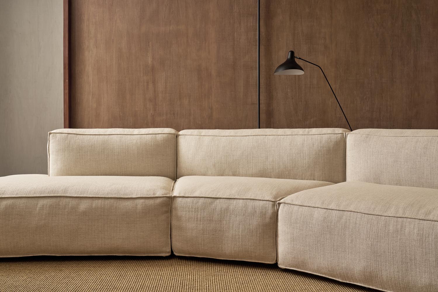 The TEST Baker Inline Sofa by Wooden Stories, featuring a modern beige modular design and clean lines, sits against a brown wooden wall with a black wall-mounted lamp above and a neutral textured rug underfoot.